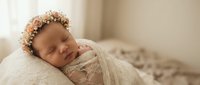 Peaceful newborn baby girl sleeping wrapped in soft cream blanket wearing delicate floral headband in warm natural light studio portrait for nursery decor.