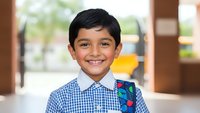 Happy young South Asian boy student in checkered school uniform shirt smiling at camera in classroom setting for educational content and back to school materials.