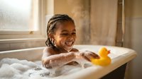 Young African American girl enjoying bubble bath with rubber duck toy in bright bathroom with natural window light.