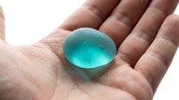Caucasian hand holding a translucent teal water gel ball with tiny bubbles, isolated on white background.