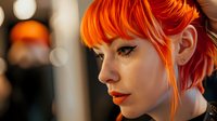 Young woman with vibrant orange hair and dramatic makeup in profile view against blurred warm background, showing modern alternative style and bold fashion choices.