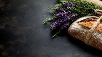 Rustic artisan bread loaf tied with twine alongside fresh purple lavender sprigs on dark moody background, food photography composition with copy space.