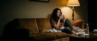 Sick woman with dark curly hair sitting on brown couch holding tissues, surrounded by used tissues, with lamp and bottle nearby, depicting cold or flu symptoms.