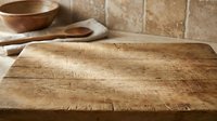 Rustic wooden cutting board on kitchen counter with natural wood grain texture and warm lighting for culinary food preparation and cooking backgrounds.