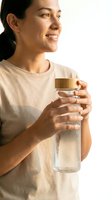 Smiling young Hispanic woman holding a clear reusable water bottle with bamboo lid, wearing a beige t-shirt, healthy hydration lifestyle concept.