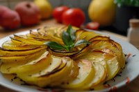 Golden roasted potato slices arranged in elegant spiral pattern with fresh herbs on white plate, surrounded by colorful vegetables in rustic kitchen setting.