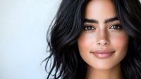 Young woman with dark wavy hair and natural makeup smiling confidently against light background. Soft beauty portrait showcasing glowing skin and gentle expression.