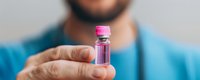 Medical professional in blue scrubs holding small glass vial with pink liquid medicine against blurred background, demonstrating pharmaceutical sample or vaccine dose.