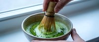 Hands whisking matcha powder into hot water with traditional bamboo brush in ceramic bowl, creating frothy green tea beverage for Japanese tea ceremony preparation.