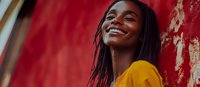 Young African American woman with dreadlocks wearing yellow top smiling genuinely against vibrant red wall background, expressing joy and confidence in casual outdoor setting.
