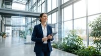 Professional businesswoman in navy suit holding tablet in modern glass office building with natural light and plants creating corporate workspace atmosphere.