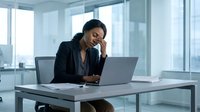 Stressed African American businesswoman experiencing headache while working on laptop in modern office environment with large windows and natural light.