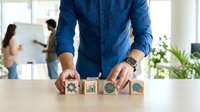 Man in blue checkered shirt arranging wooden blocks with business icons on white table, colleagues working in background office environment, representing strategy and planning.
