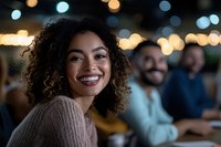 Young African American woman with curly hair and bright smile at evening social gathering, warm bokeh lights create festive atmosphere in background.
