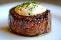 Grilled beef tenderloin filet topped with creamy butter sauce and fresh chopped herbs on white plate, close-up view showing juicy meat texture and caramelized crust.