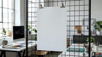 Modern office workspace with blank white poster mockup hanging on black metal grid wall, laptop on desk, industrial interior design for branding presentation.