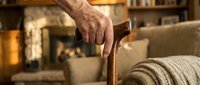 Elderly person's wrinkled hand gripping wooden walking cane in cozy living room with fireplace, representing aging, mobility assistance, and home comfort.