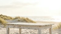Weathered wooden table on sandy beach with coastal dunes and ocean in background, suitable for lifestyle and travel photography.