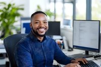 Cheerful African American male professional in navy blue shirt working at computer desk in modern bright office space, displaying genuine smile and confidence.