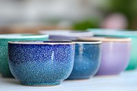 Colorful ceramic bowls with speckled glaze finish arranged in row showing gradient of blue, purple and turquoise hues, selective focus on handcrafted pottery.