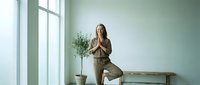 Young Caucasian woman practicing yoga tree pose in minimalist green room with plant and natural light for wellness and mindfulness meditation.