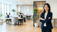 Confident female professional with crossed arms standing in modern office lobby, wearing dark blazer, with colleagues working at desks and green plants in background.