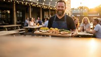 Happy bearded chef serving grilled food on wooden board at outdoor restaurant patio during golden hour sunset with string lights and dining guests.