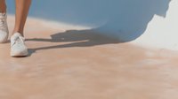 Person in white sneakers walking on sandy beach surface with soft shadow cast against light blue wall, creating minimalist summer aesthetic.