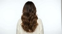 Young woman with long wavy brown hair from behind wearing cream knit sweater against white background for hair care beauty salon advertising.