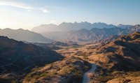 Winding mountain road through dramatic desert landscape with rugged peaks and golden valleys at sunset, aerial perspective capturing remote wilderness terrain.