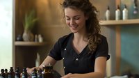 Young female massage therapist in black uniform arranging essential oils and preparing workspace in spa salon with warm ambient lighting and modern decor.