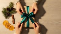 Hands wrapping Christmas gift with teal ribbon bow on wooden table with festive decorations including pine branches, cinnamon sticks and dried oranges.