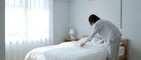 Professional housekeeper making bed in modern hotel room with white linens and natural lighting for hospitality service industry.