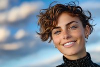 Young woman with curly brown hair and bright smile against blue sky background. Natural portrait for lifestyle and wellness campaigns.