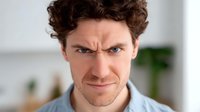 Caucasian male with curly dark hair expressing skeptical and disapproving emotion, wearing light blue shirt against blurred home interior background.