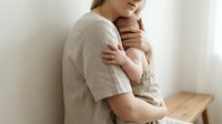 Young Caucasian mother gently holds her newborn baby against her chest, sharing a tender bonding moment in a soft, minimalist interior.