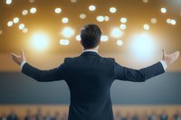 Businessman in dark suit spreading arms wide on stage with blurred bokeh lights and audience background, view from behind, success concept.