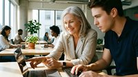 Mature woman with gray hair and younger man collaborating on laptop in modern office workspace with colleagues working in background, natural lighting from windows, professional mentoring and teamwork concept.