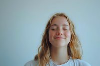 Young Caucasian woman with blonde wavy hair and closed eyes expressing peaceful contentment while wearing white t-shirt against light blue background.