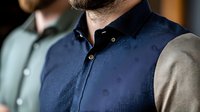 Professional man wearing navy blue business vest with beige sleeves and button-down collar, cropped view showing torso and neck area against blurred background.