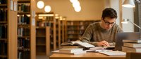 Young Caucasian male student with glasses studying at library desk with open books and laptop computer surrounded by warm lighting and bookshelves.