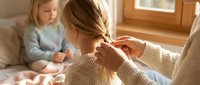 Mother braiding young daughter's hair in cozy bedroom while toddler watches from bed. Warm family bonding moment showcasing maternal care and sibling connection.
