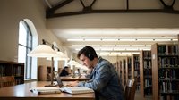 Young Asian male student studying with laptop and books in quiet university library with wooden tables and bookshelves in background.