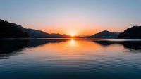 Tranquil lake at sunrise with golden sun reflecting on calm water surface surrounded by mountain silhouettes and forest creating peaceful morning atmosphere.