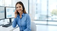 Smiling young Caucasian businesswoman with headset working at modern office desk with computer monitors displaying charts and data analytics.