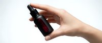 Female hands holding dark amber glass dropper bottle with black pump dispenser against clean white background for skincare and cosmetic product presentation.
