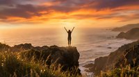 Woman with raised arms celebrating freedom on coastal cliff at golden sunset overlooking ocean waves and dramatic sky for wellness and achievement concepts.