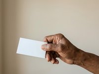 Senior male hand with medium brown skin tone holding blank white card against neutral beige background for business, identity, or promotional use.