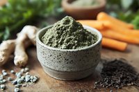 Green superfood powder in ceramic bowl with fresh ginger root, carrots and spirulina ingredients on rustic wooden background. Natural dietary supplement.