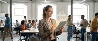Young woman with brown hair and medium complexion wearing a tan blazer holds a tablet while smiling in a modern, bright office space with diverse colleagues working collaboratively in the background.
